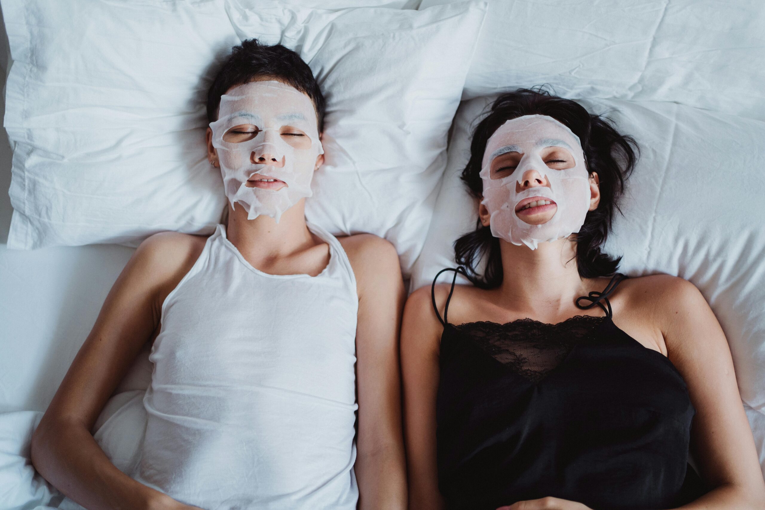 A couple lies in bed with sheet masks, promoting skincare and relaxation.